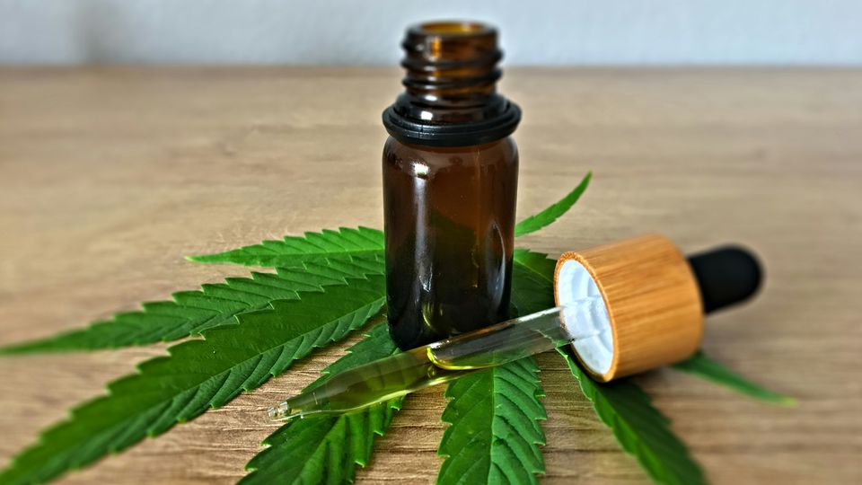 A bottle of CBD and a pipette on top of a cannabis leave on a wooden desk.