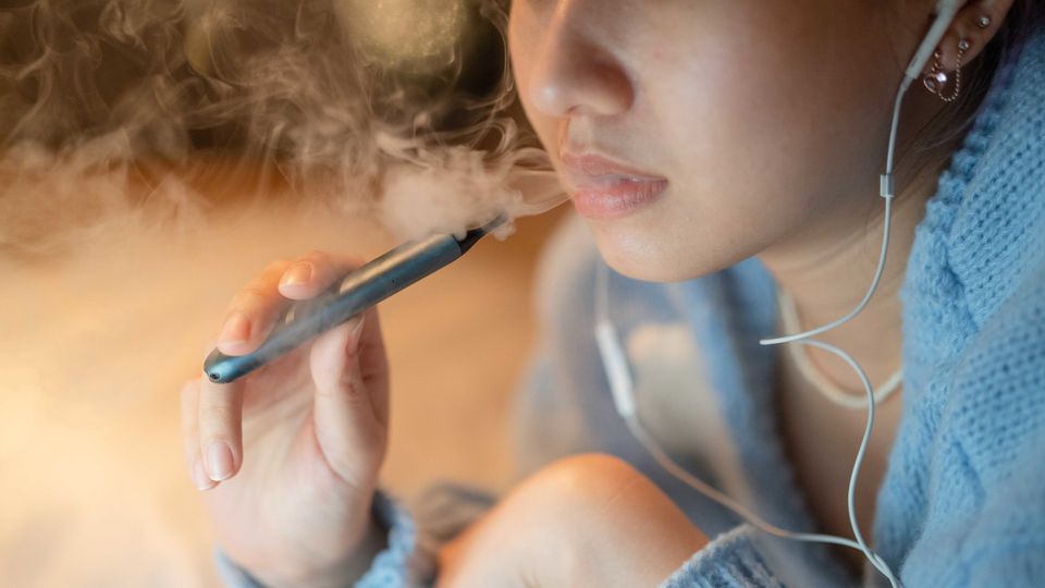 Young woman vaping an e-cigarette while wearing earbuds indoors.