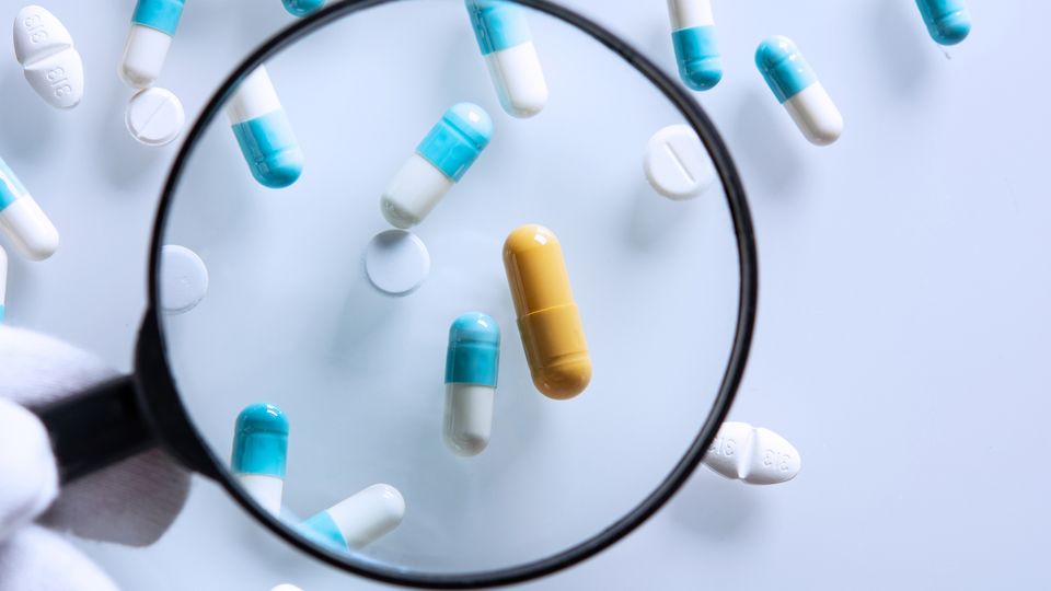 A variety of different pills on a bench with one yellow pill under a microscope, representing drug repurposing.