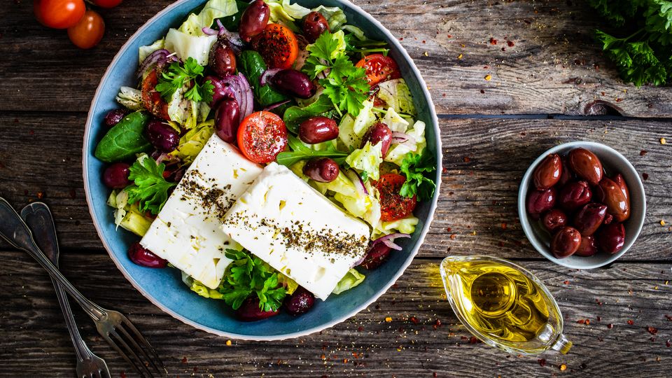 A bowl of greens, tomatoes, olives and cheese on a wooden table. Cutlery, olive oil and more olives also present.
