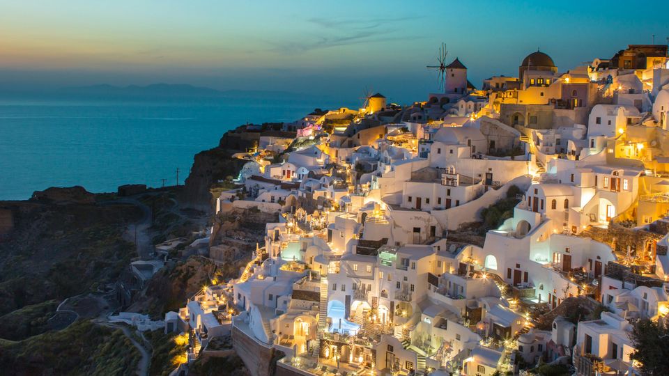 A sunset view of Santorini, white houses in the hills, blue sea in the background.
