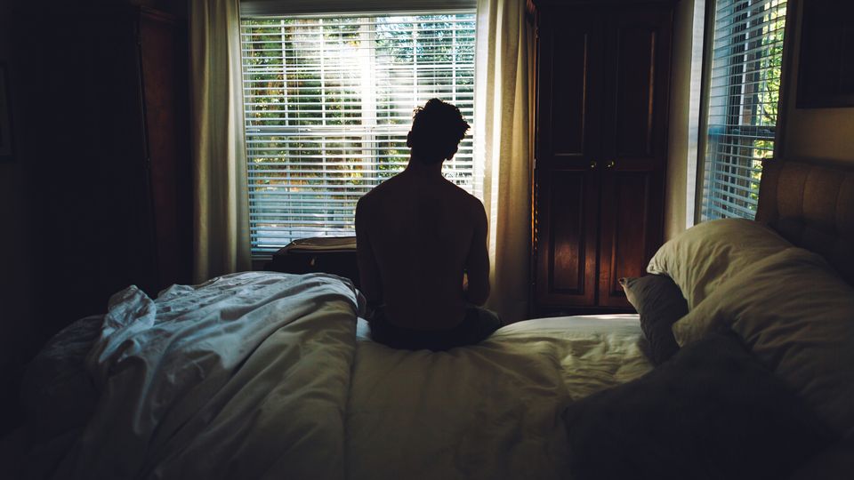 A man suffering with depression sat on his bed in the dark in front of a window.