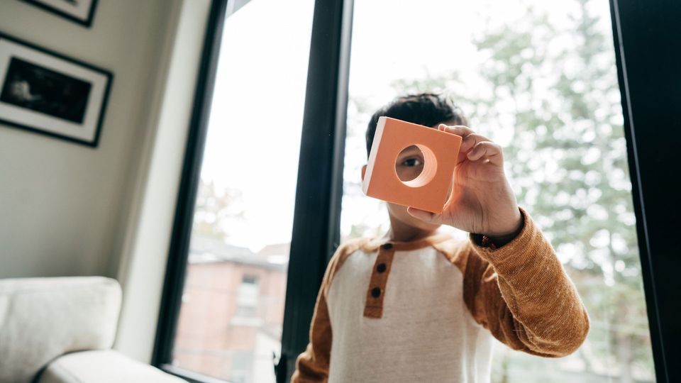 Child holding an orange toy brick with a circle cut out of it, up to his eye.