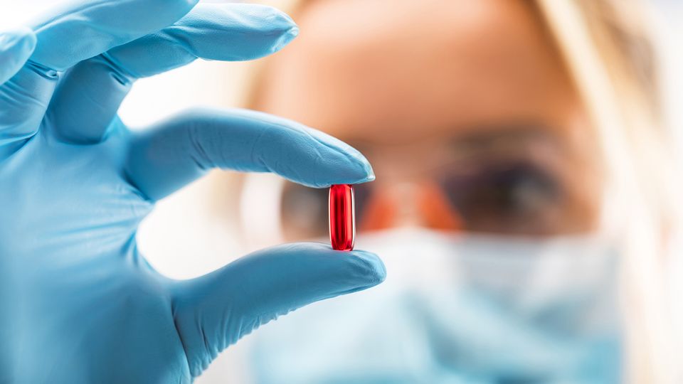 A female scientist in gloves and a mask holding up a red pill, representing drug discovery.