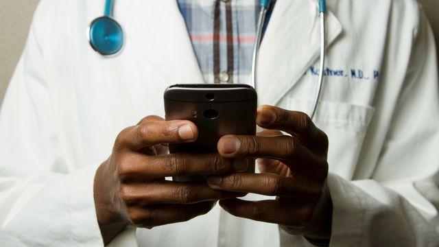 A person in a doctor's coat (with stethoscope around their neck) looks at their phone. Their face is not visible. 