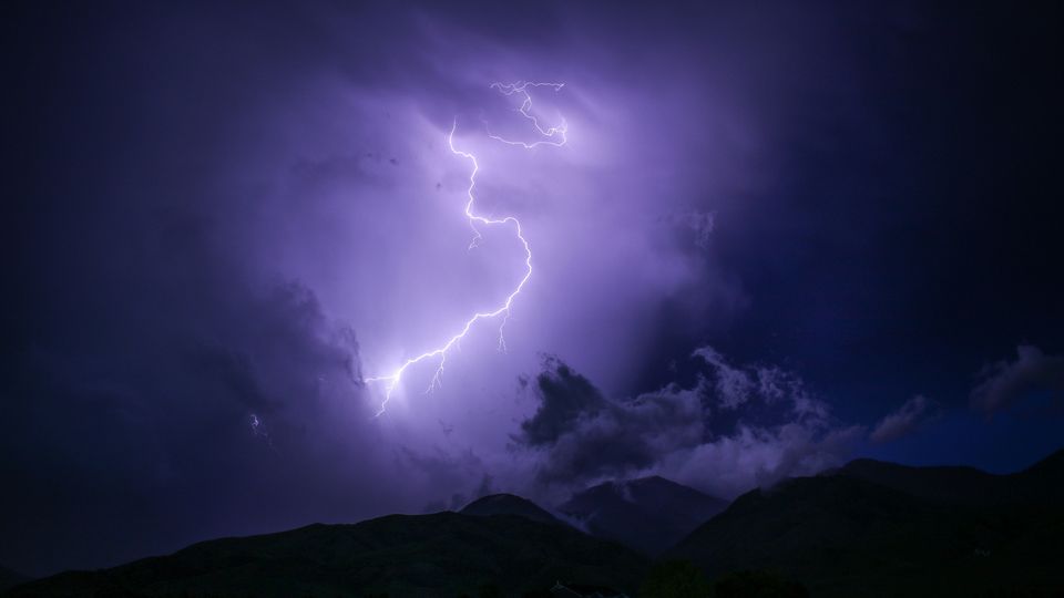 A streak of purple lightning in the sky above the silhouettes of some black hills.