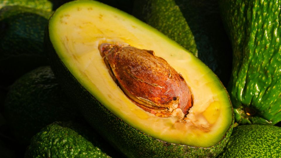 A halved avocado, its brown stone and yellow flesh visible, on a pile of uncut green avocadoes.