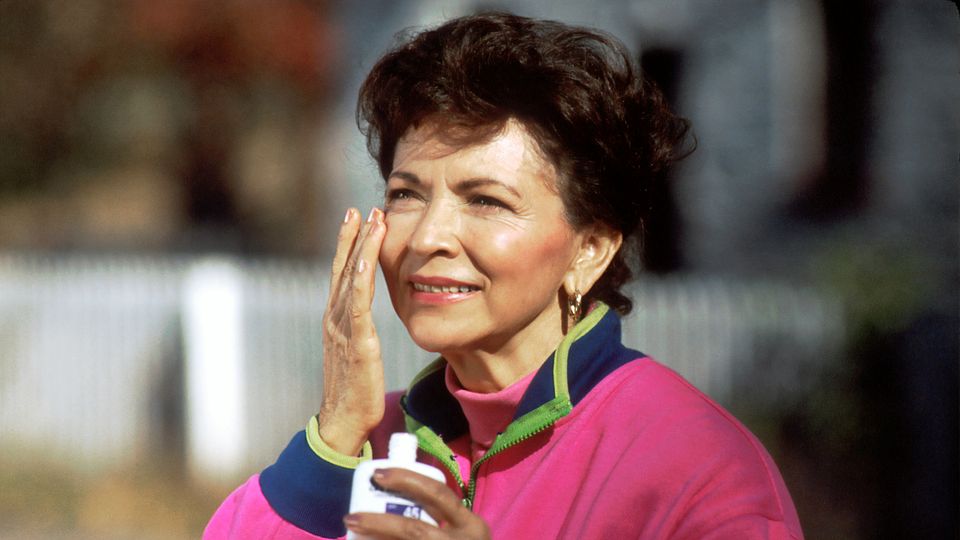 A person in a pink jacket applies sunscreen to their cheek.