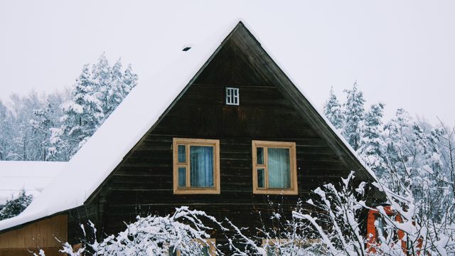 A photograph of a wooden house with a triangular roof and large windows, covered by a blanket of snow and surrounded by snowy trees. 