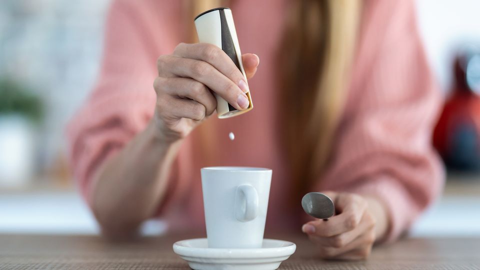 Person with long blond hair in pink jumper adding sweetener to their white mug. Spoon in hand.