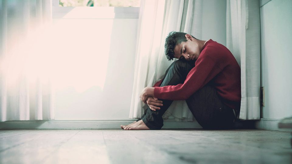 Young man sitting alone on the floor by a window, hugging his knees in a posture of loneliness.
