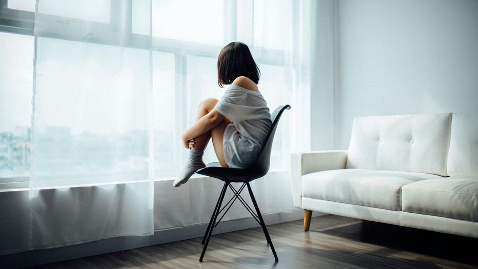 Young woman sitting alone on a chair by the window, reflecting symptoms of depression.
