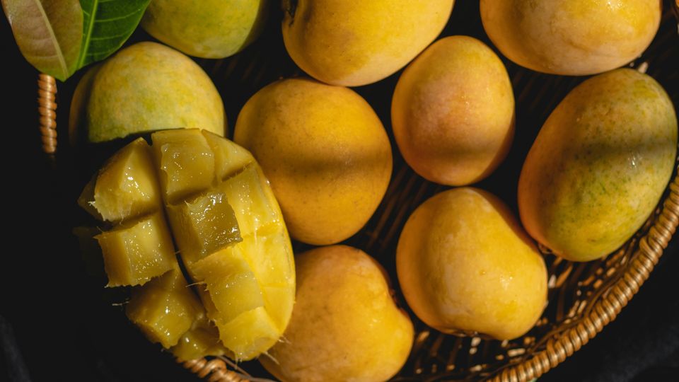 A woven basket filled with yellow mangoes, one already cut.