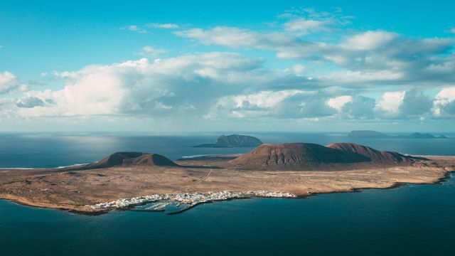 A helicopter/drone shot of one of the Canary Islands. Blue sky, blue sea, mountains and town visible.
