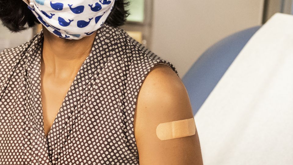 A South Asian woman in a brown sleeveless top and white and blue face mask, with a band-aid on her upper arm.