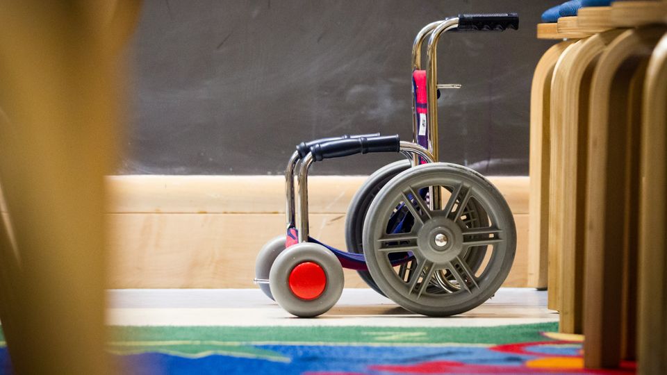 Pediatric wheelchair beside chairs in a classroom, symbolizing mobility challenges in children with alternating hemiplegia of childhood.