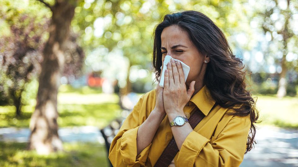 Woman in a yellow jacket sneezing into a tissue while walking in a park.