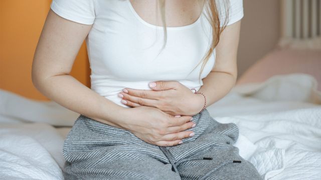 Woman sat on edge of a bed holding her stomach in pain.