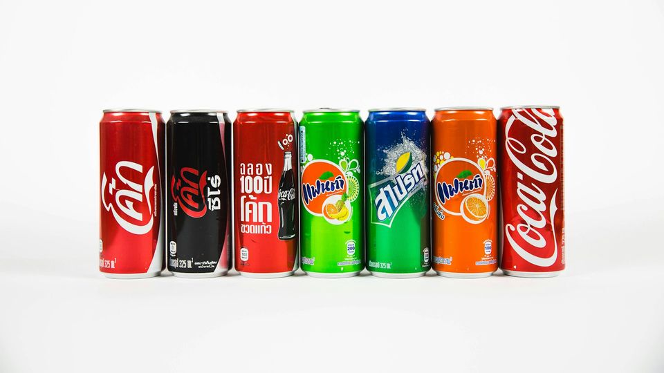 Assorted soda cans including Coca-Cola, Fanta and Sprite flavors lined up against white background.