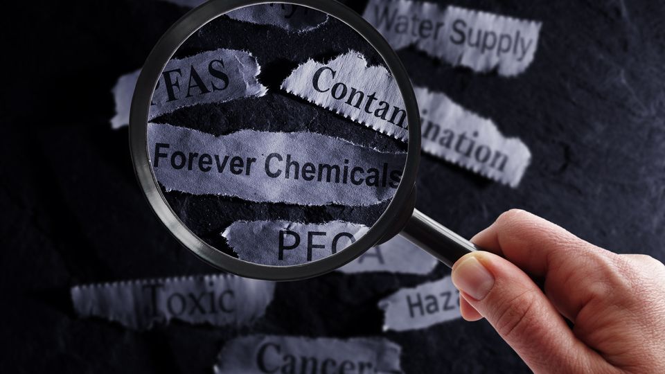 A person holds a magnifying glass over several strips of paper that read "forever chemicals", "PFAS", "Contamination" and other related words.
