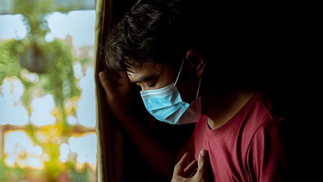 Man wearing a face mask looking out window with hand on chest, symbolizing COVID-19 distress.