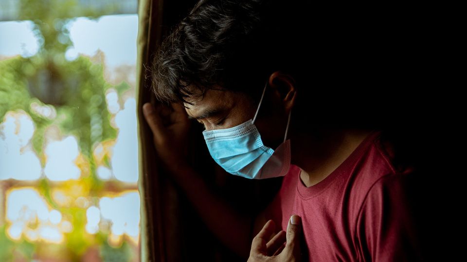 Man wearing a face mask looking out window with hand on chest, symbolizing COVID-19 distress.