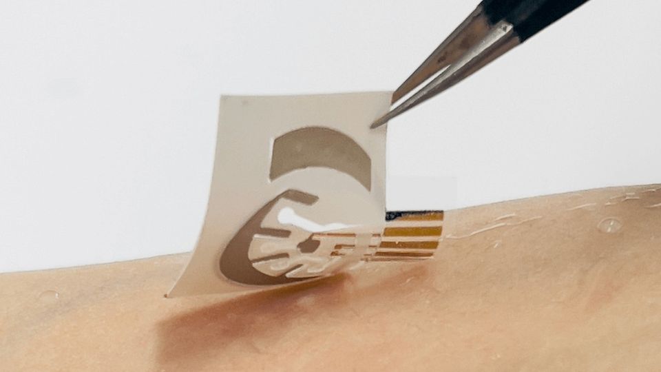 A small sticker with printed silver electronic circuits being placed on a person's arm using a pair of tweezers.