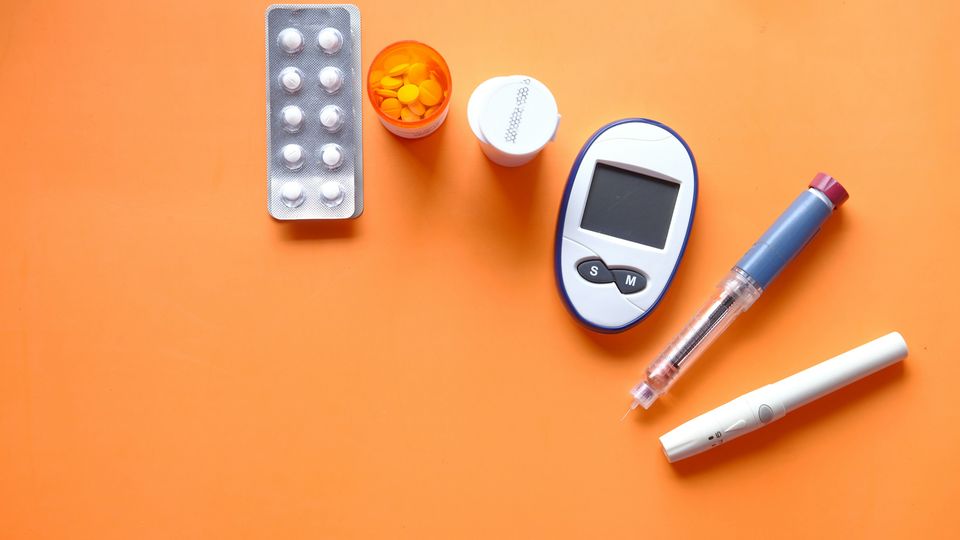 A blister packet and pill pot containing diabetes drugs next to a glucose monitor and an insulin pen on an orange background.