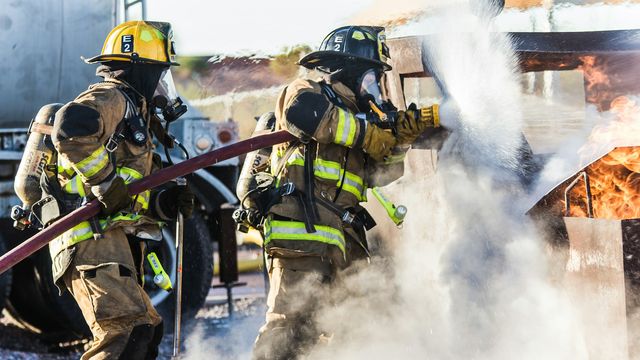 Firefighters in full gear using a hose to extinguish a burning structure during an emergency response.