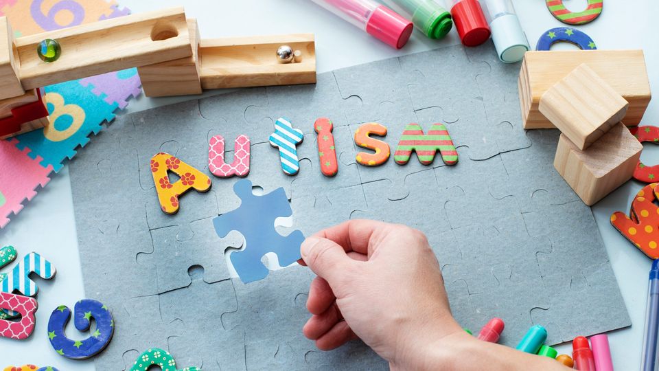 Hand placing a puzzle piece beneath colorful letters spelling “autism” surrounded by toys.
