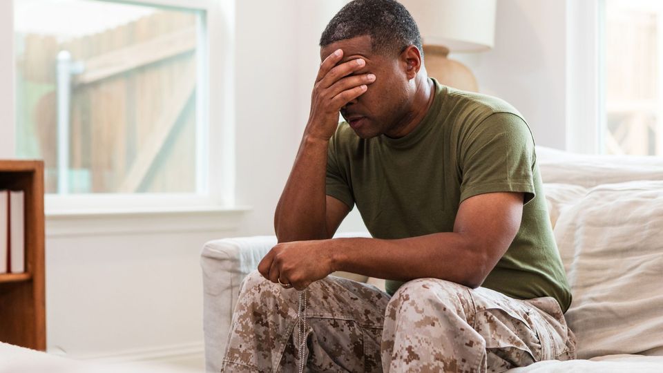 Worried military veteran in uniform sitting on a couch with head in hand, symbolizing PTSD.