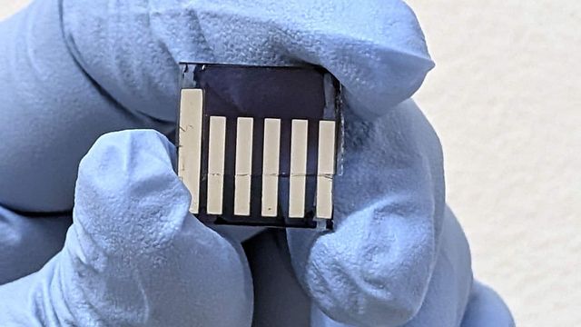 A person wearing blue laboratory gloves holding a small, clear, square chip. On it are six small metal bars. 