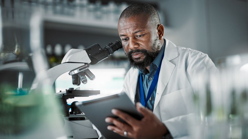 Scientist in a lab coat analyzing data on a tablet beside a microscope in a research laboratory.