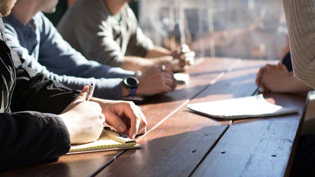 A group of people sat around a wooden bench, notepads and pens visible. 