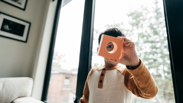 A child stood in front of a window holding up a block toy. 