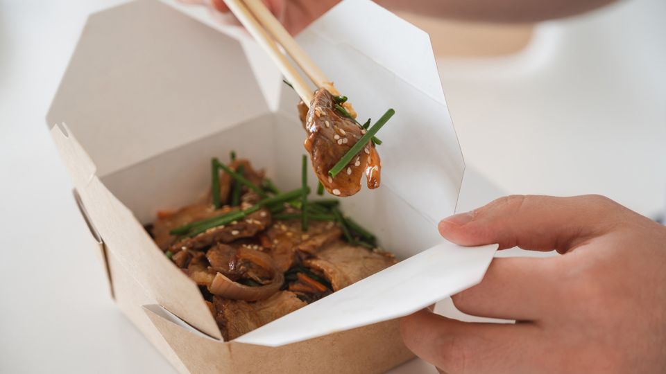 Hand holding a cardboard takeaway box of food and another using chopsticks to pick the food up to eat.