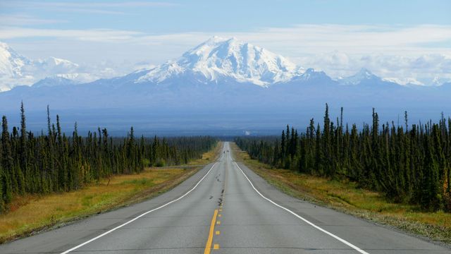 A long road into the horizon, lined with coniferous trees, an Alaskan mountain the background. 