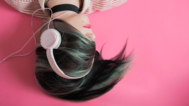 Person with black and green hair wearing white headphones lying on a pink background.
