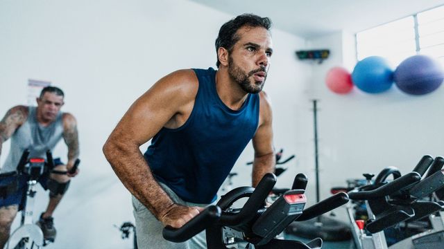 Focused male athlete in a blue tank top riding a stationary bike in a gym. 