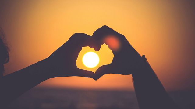 Two people's hands create a heart shape in front of a sunset, symbolizing connection and love. 