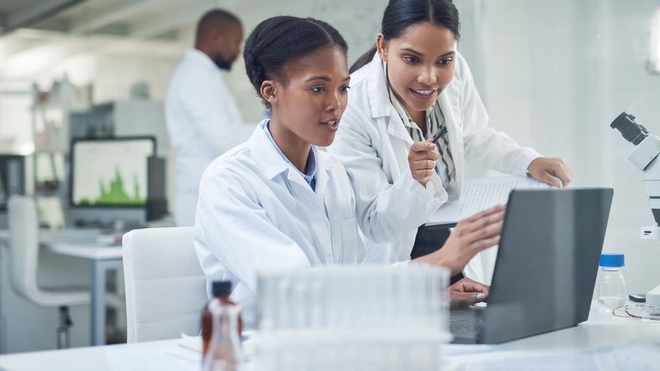 Two scientists in lab coats collaborating on research while reviewing data on a laptop in a laboratory.