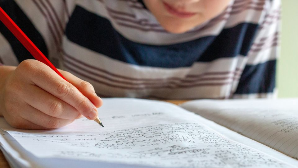 A young boy in a striped shirt writing in an open notebook with a pencil.