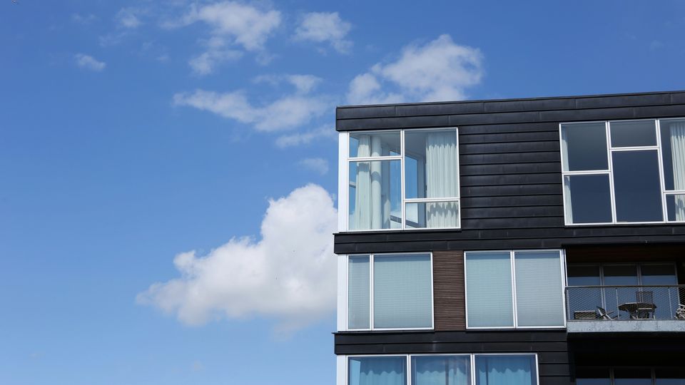 The top corner of an apartment block, featuring many apartment windows, against a light blue sky.