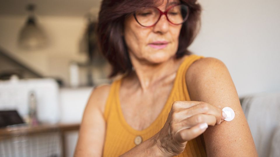 Woman applying a hormone therapy patch on her upper arm at home.