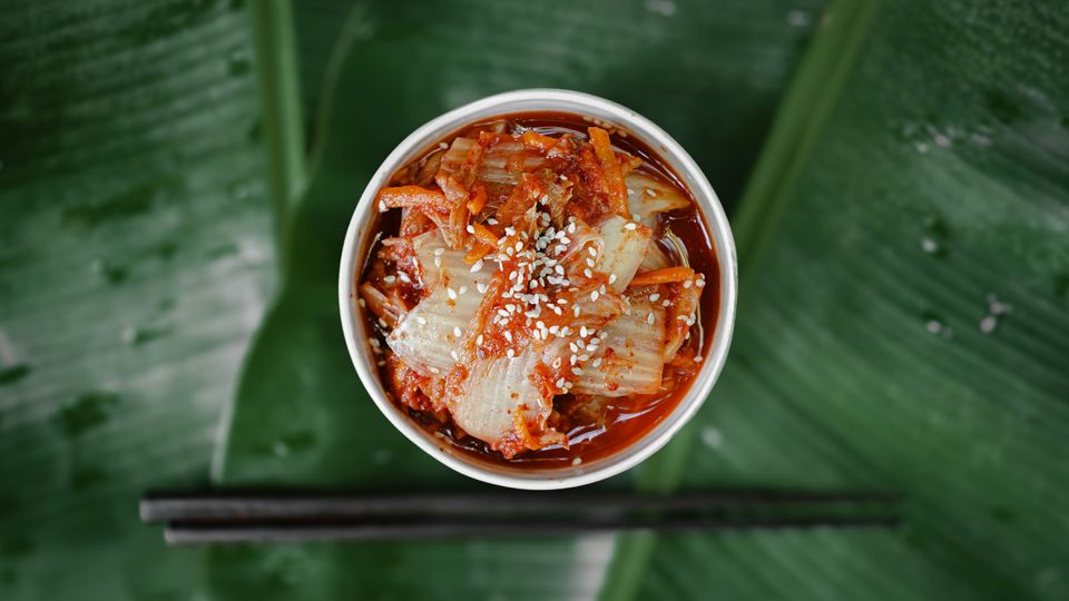 A bowl of kimchi cabbage, red orange and pale yellow, shot from above. Green leaves and black chopsticks visible in background.