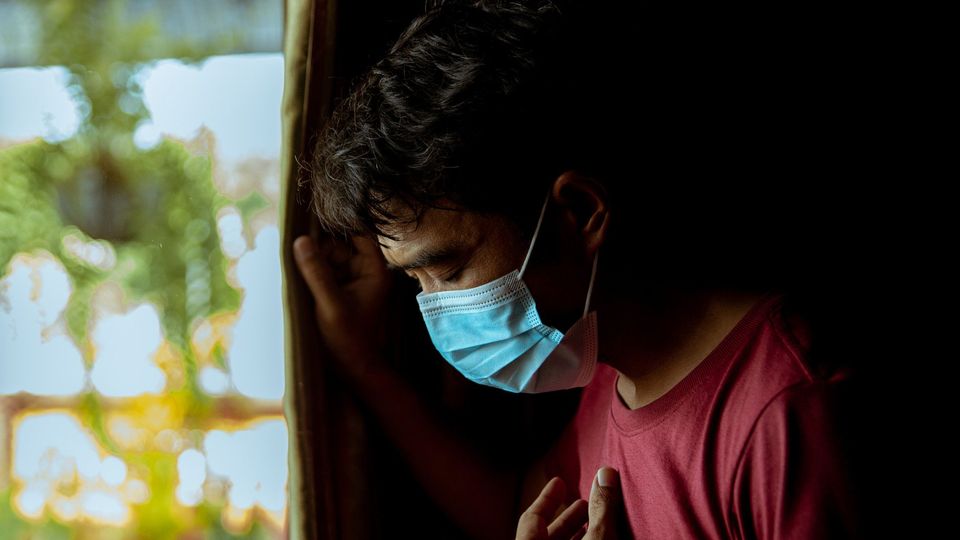 Man wearing a face mask looking out the window, symbolizing the impact of COVID-19 and isolation.