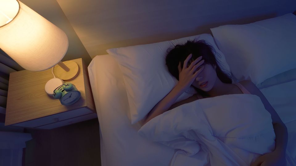 Woman lying in bed with her hand on her forehead, showing discomfort from irregular sleep patterns.