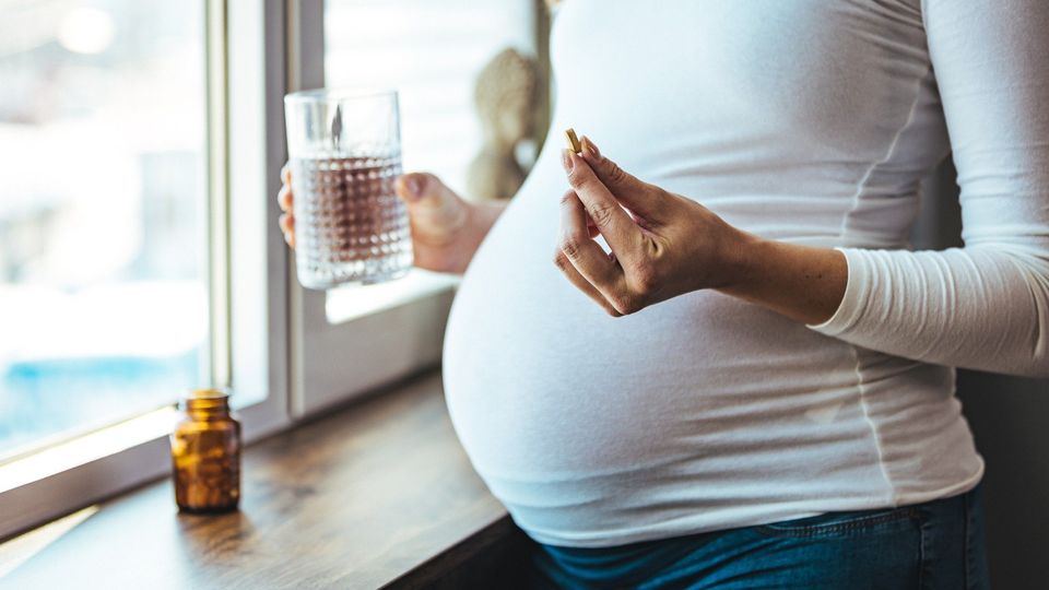 Pregnant woman holding prenatal vitamins and a glass of water by a window.