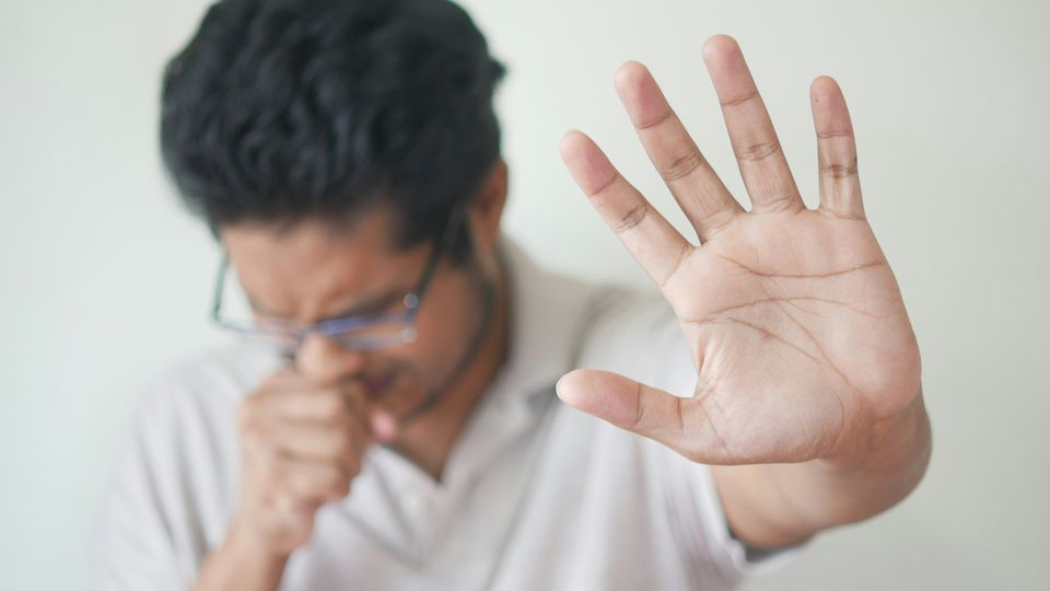 A man with his head down, coughing into his fist, with an outstretched hand in a "stop" gesture being shown towards the camera.