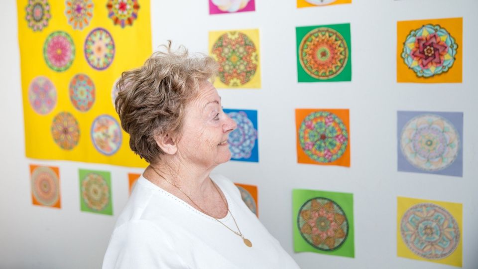 Smiling older woman looking at colorful mandala artwork displayed on a wall.
